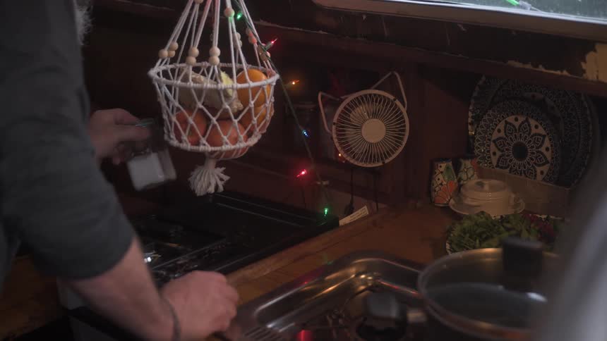 A man retrieves a jar of spices from a cabinet in a vibrant kitchen adorned with colorful string lights a hanging fruit basket and located on a boat
