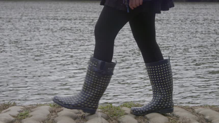 A woman wearing rain boots is walking along a path next to a river