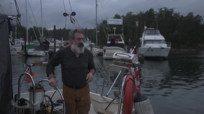 A bearded man on a boat pulls a rope while other boats and a dock are visible in the cloudy background