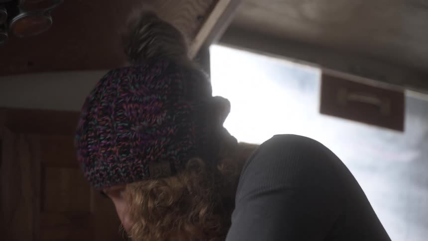 A blonde haired woman wearing a colorful beanie works in a small cramped and cozy home kitchen