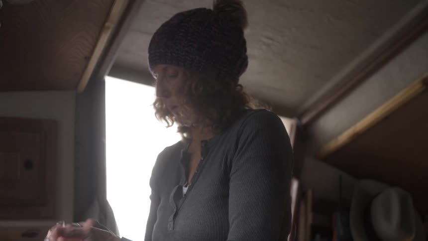 A blondehaired woman wearing a colorful beanie works in a small cramped and cozy home kitchen