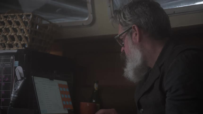 A bearded man with glasses works on a laptop in a dimly lit room pausing to wipe his nose with a tissue