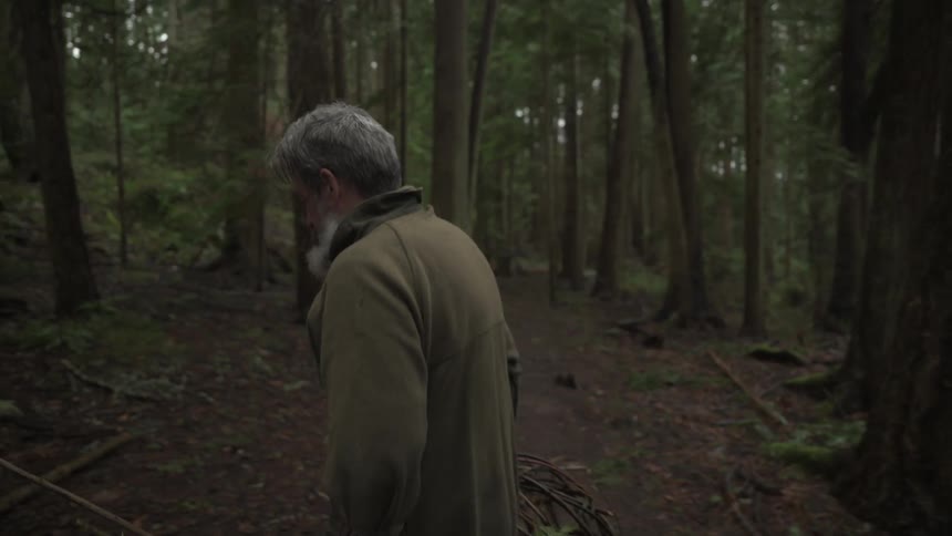 A man drops a bundle of sticks and pulls out his knife