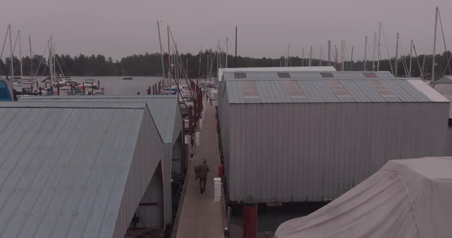 An aerial shot follows a person walking along a dock through a marina filled with covered boat slips and numerous boats under an overcast sky