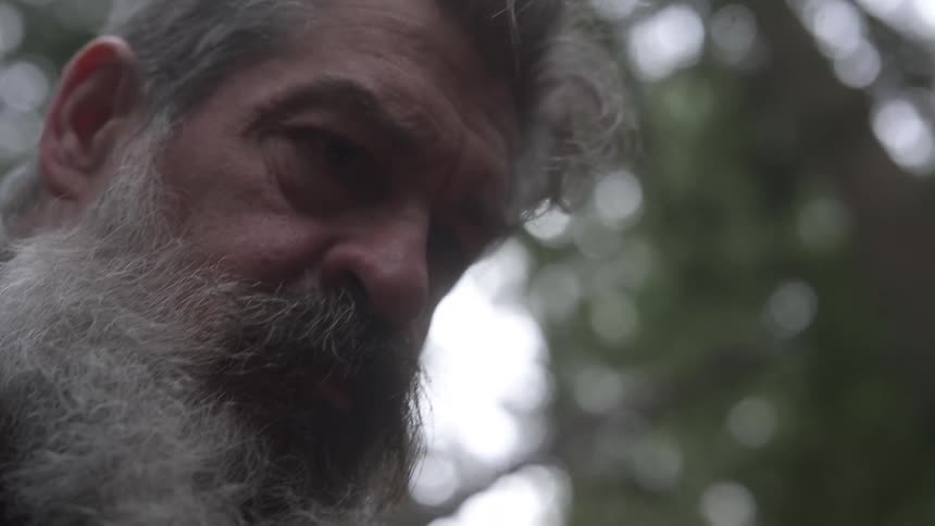 Older bearded man grimaces and gestures while examining something in the forest during the daytime