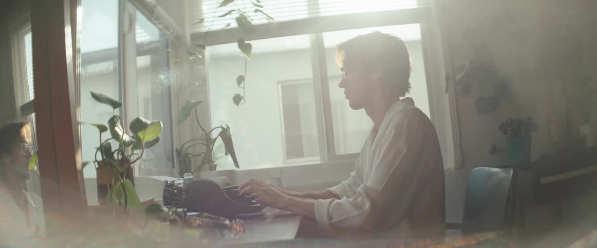 A man sits at a desk by a bright window focused on typing on a vintage typewriter surrounded by plants