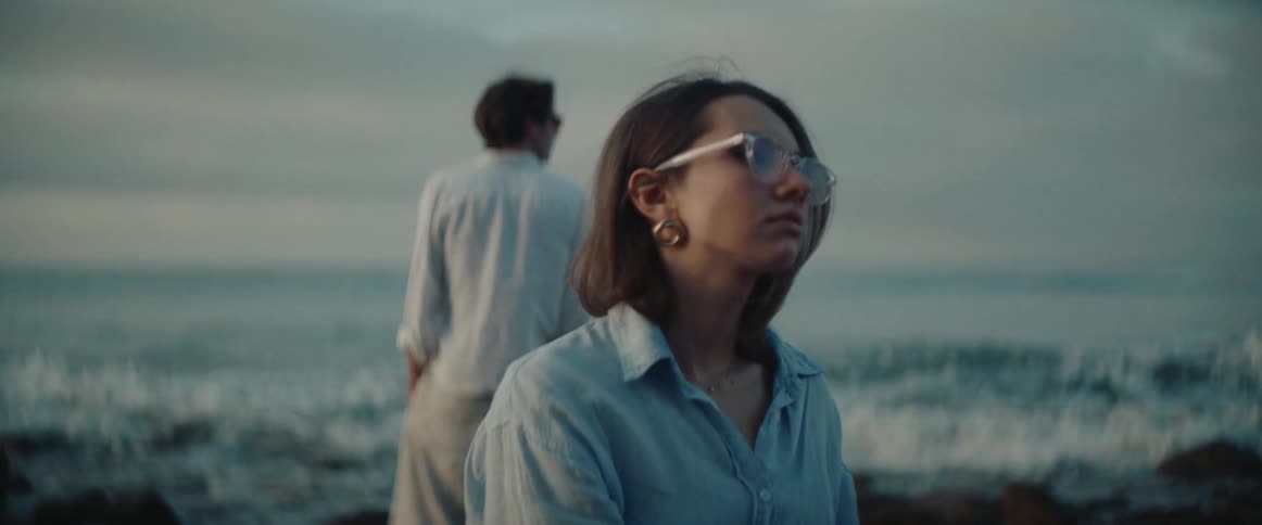The camera focus shifts between a woman in the foreground and a man in the background both standing near the ocean shoreline facing opposite directions