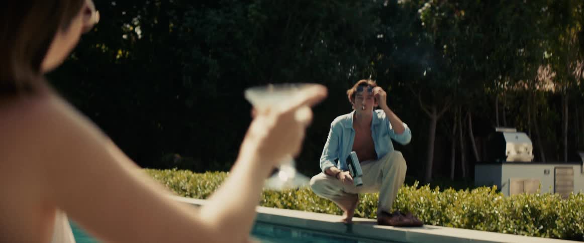 A photographer crouches next to a swimming pool and films a woman holding a martini