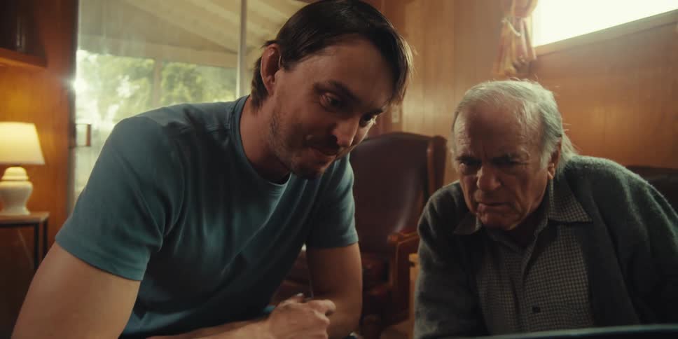 A young man sits beside an elderly man in a cozy home on a sunny day both focused on a laptop