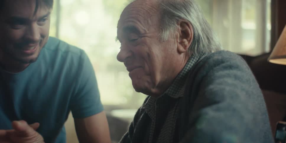 A young man sits next to an elderly man in a cozy home on a sunny day listening as the elderly man speaks with amusement
