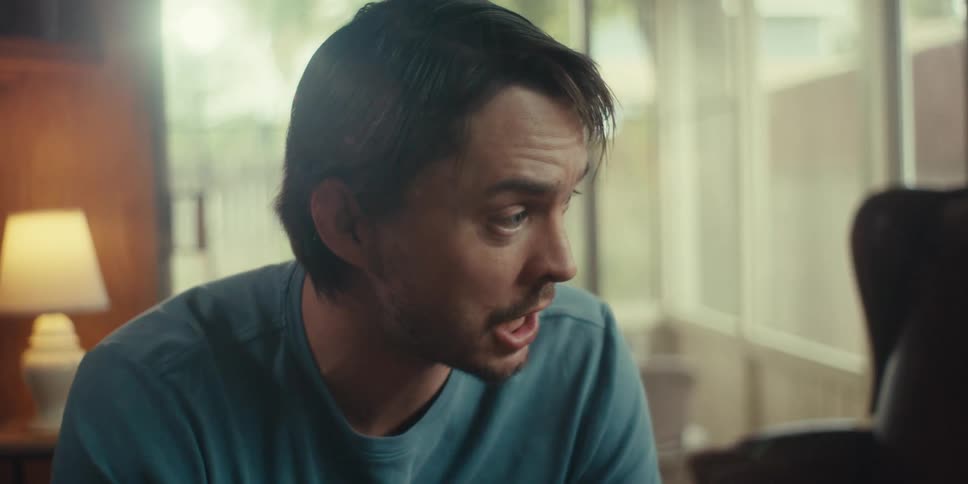 A man in a blue t shirt talks animatedly while gesturing indoors