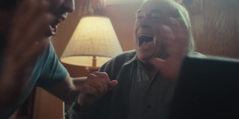 A young man and an elderly man sit together in a cozy home on a sunny day happily laughing and celebrating while focused on a laptop
