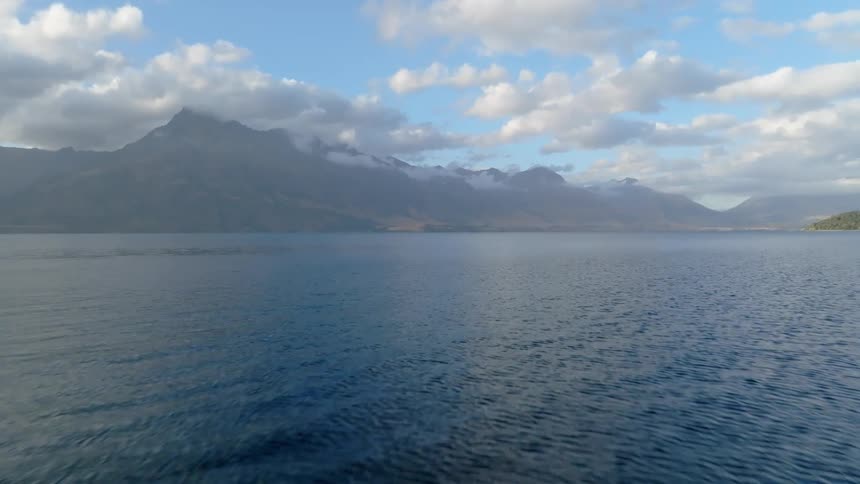 A wide view captures ripples moving across a large blue lake surrounded by hazy mountains under a partly cloudy sky
