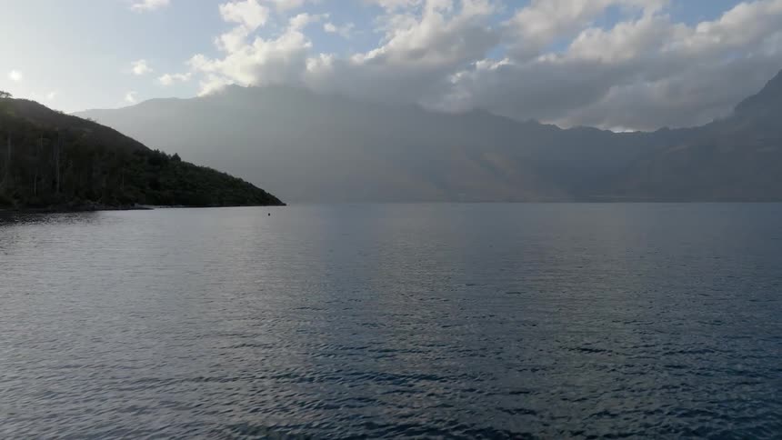 A vast calm lake stretches towards a dark tree covered hillside and distant hazy mountains under a partly cloudy sky