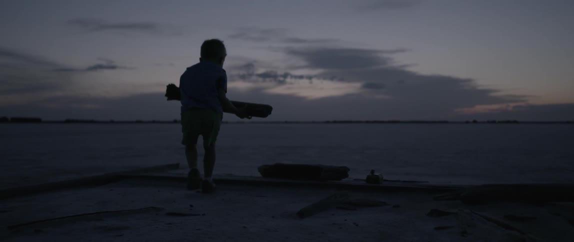 A boy carries a log and puts it down at dusk
