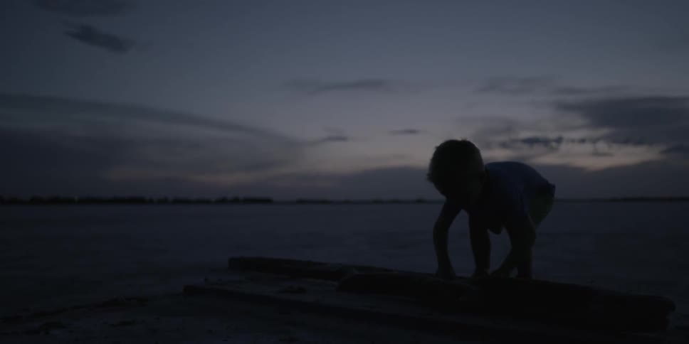 A young boy lifts a large stick in an open area at dusk