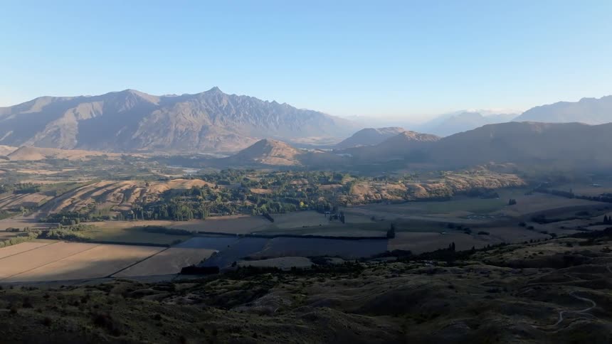 A vast undulating landscape of hills and valleys stretches towards a hazy horizon of towering mountains under a clear pale sky
