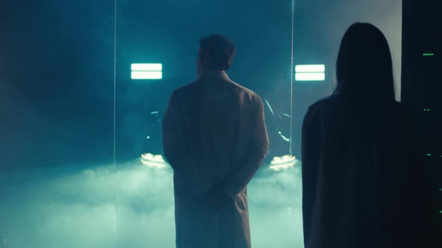 Two individuals in white lab coats stand in a dimly lit room gazing through a misted window at a car enveloped in smoke