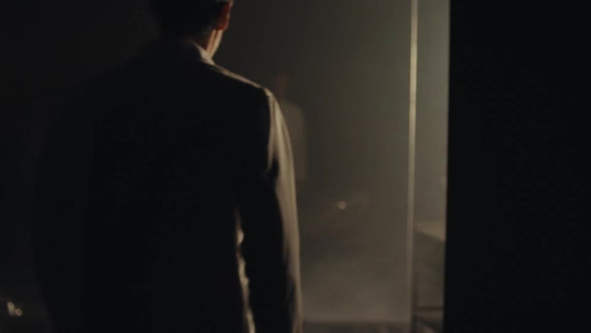A man walks through fog and stands in front of a car as the headlights turn on