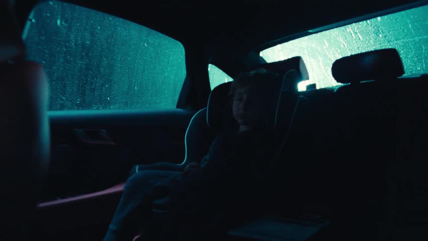 A young child sits in a car seat inside a dimly lit vehicle at night as rain falls and strobing lights illuminate the outside