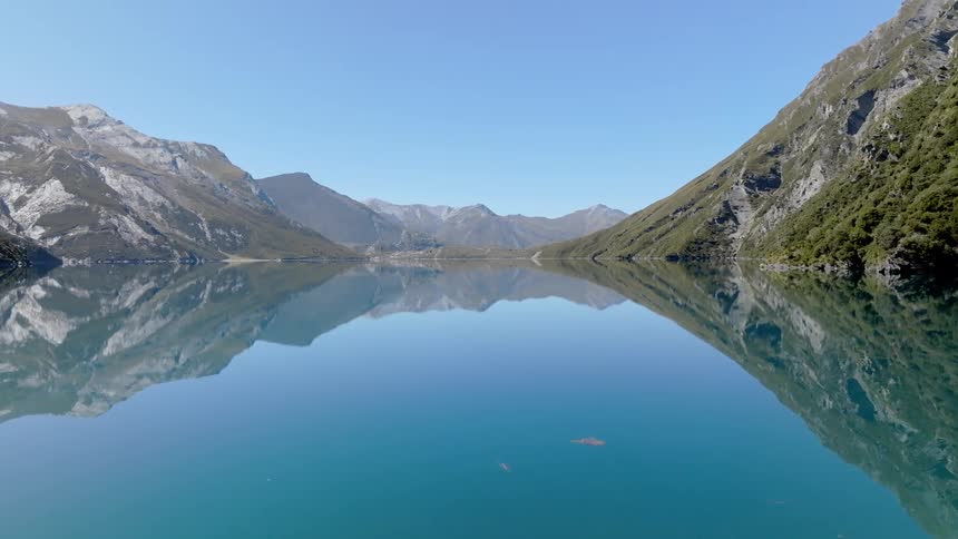 A still clear lake perfectly mirrors the steep rocky mountains that flank its edges under a bright blue sky