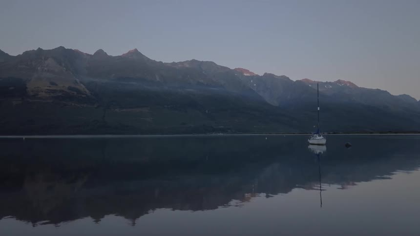 A lone sailboat rests on a calm lake reflecting the imposing dark silhouettes of a mountain range under a twilight sky