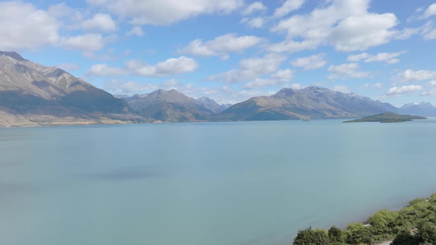 A vast pale blue lake stretches out beneath a partly cloudy sky bordered by rugged mountains and a patch of dark green trees in the foreground