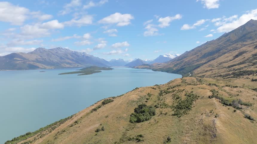 A wide pale blue lake stretches towards a backdrop of rugged mountains under a partly cloudy sky framed by dark green trees in the foreground