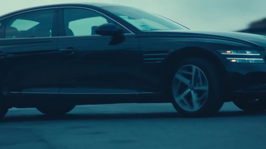 A black sedan moves along a dimly lit neighborhood road during dusk or dawn