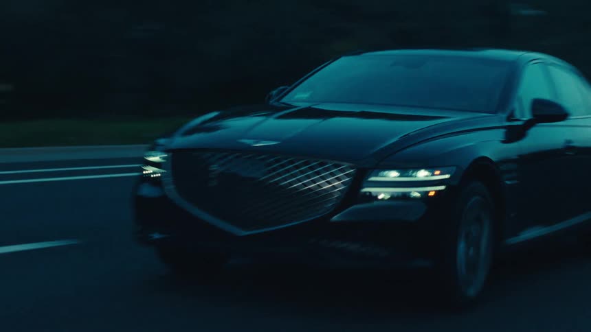 A black sedan travels along a dark road during dusk or dawn