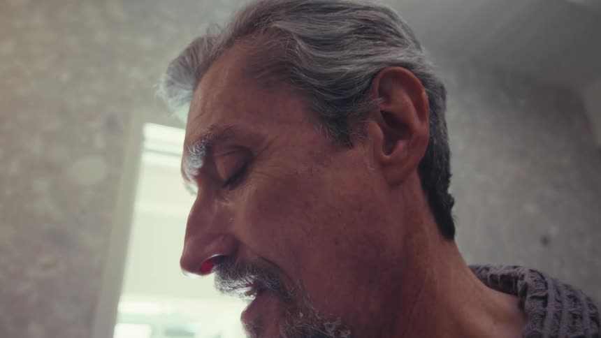 A closeup shows a person with graying hair and a beard shaving with a safety razor