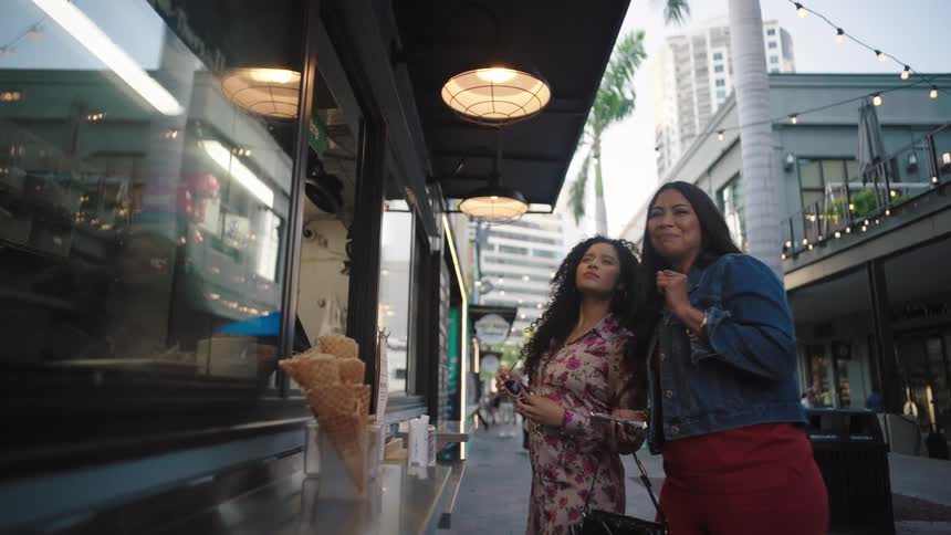 Female friends smile while standing near the outside counter of an ice cream shop and perusing the offerings