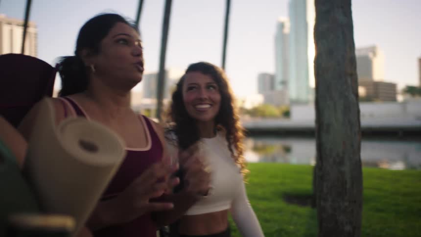 Three female friends walk through an area with grass and trees with a city behind them