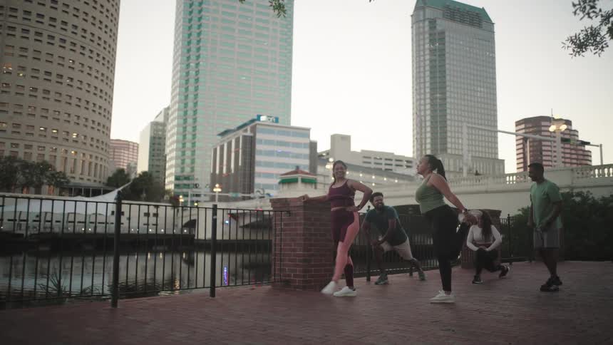 Group of people stretches along a riverbank in an urban area surrounded by skyscrapers at dusk