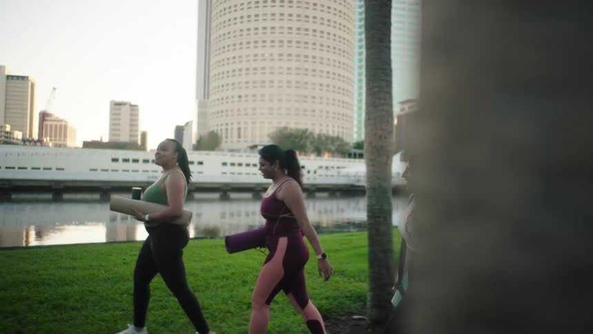 Three girlfriends walk through a park in the city holding yoga mats