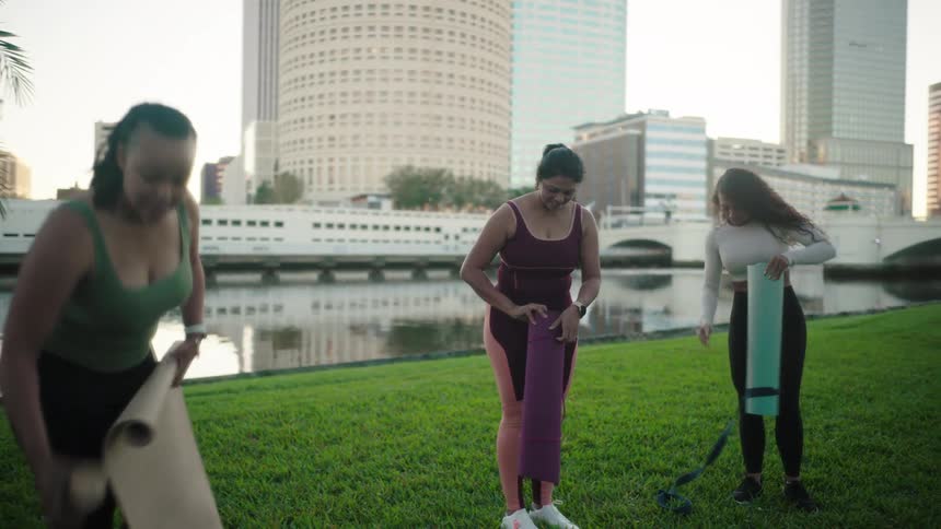 Three women begin to set up a yoga mat on a grassy outdoor area