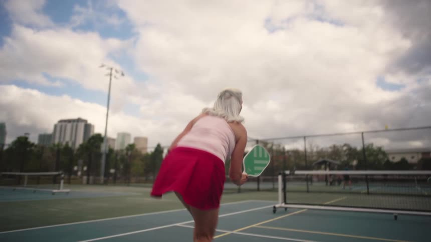 Elderly woman plays pickleball on outdoor court with cloudy sky and buildings in background