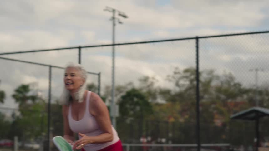 An older woman plays pickleball on a court in an urban area on a summer day