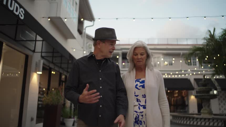 A senior couple walks toward the camera while talking and gesturing in an outdoor shopping courtyard decorated with string lights at dusk