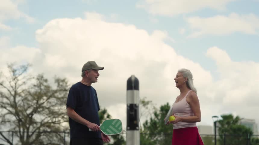 Mature couple prepares to play pickleball outdoors holding green rackets and a yellow ball under a partly cloudy sky
