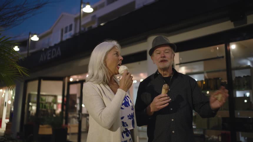 An elderly couple walk downtown in a shopping plaza while eating ice cream cones