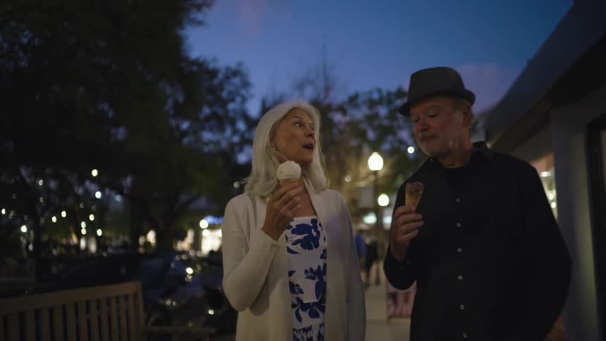 A senior couple walks along a sidewalk at dusk talking and eating ice cream cones under ambient street lights