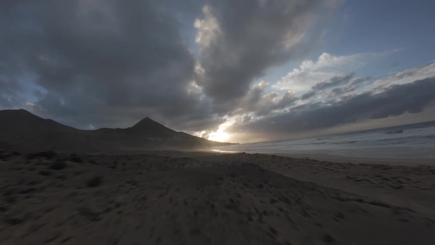 Sun shines from the mountains on the horizon onto a beach under a cloudy sky