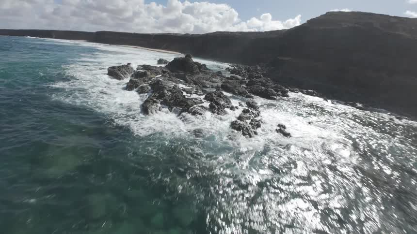 A dramatic coastline with crashing waves meeting the deep blue ocean under a partly cloudy sky
