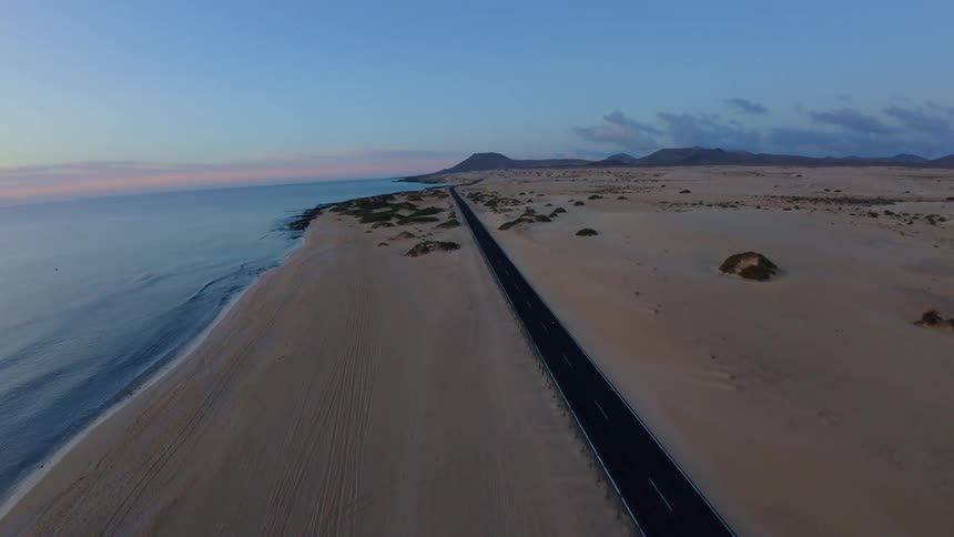 Paved road cuts through a desert landscape by the sea at dusk with mountains in the background and the sky in soft shades of blue and pink