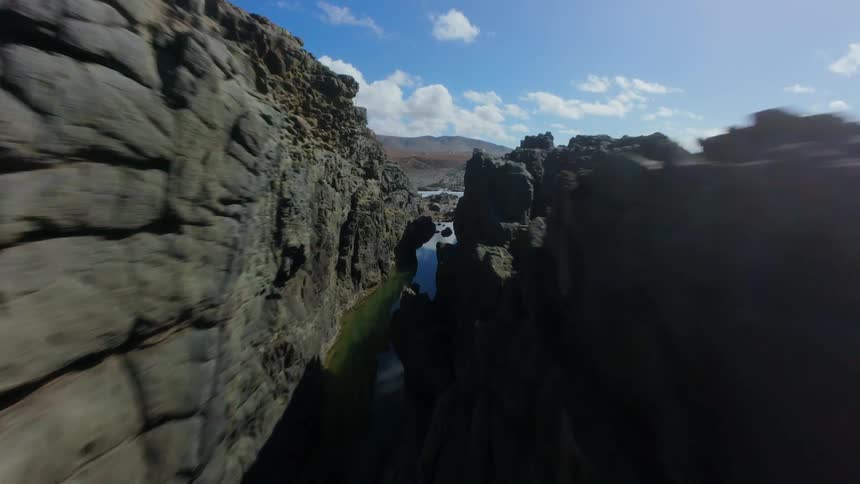 A rugged rocky coastline with dramatic cliffs meeting the ocean under a bright blue sky dotted with fluffy clouds