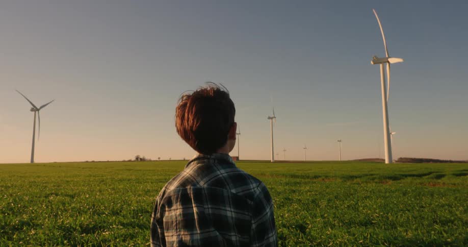 Seen from behind a young boy stands in a vibrant green field gazing towards numerous wind turbines on the horizon during sunset or sunrise