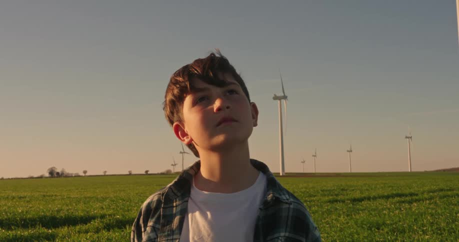 A teenage boy stands outside on a wind farm and looks up at a windmill