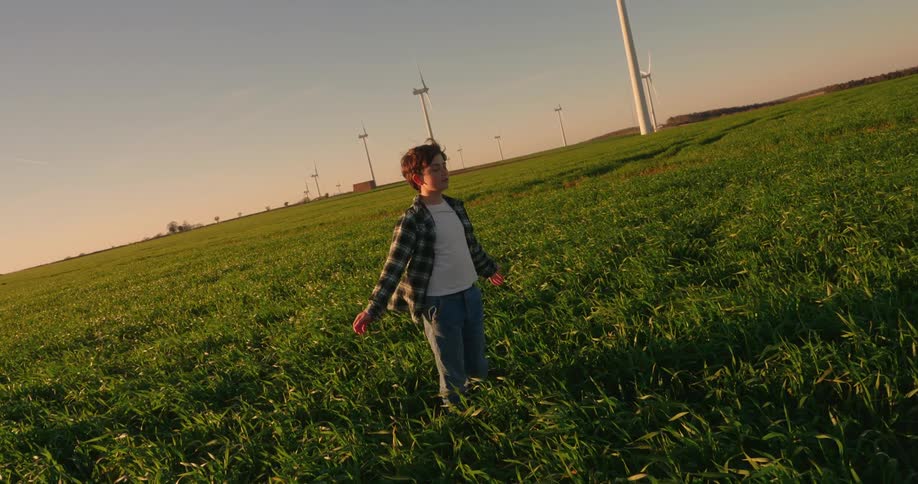 A boy stands in a green field with several wind turbines scattered across the landscape under a clear blue sky