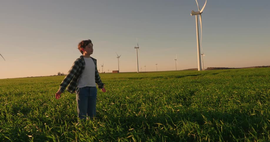 A teenage boy is standing in a long grass field with a wind turbine farm behind and he has his arms outstretched